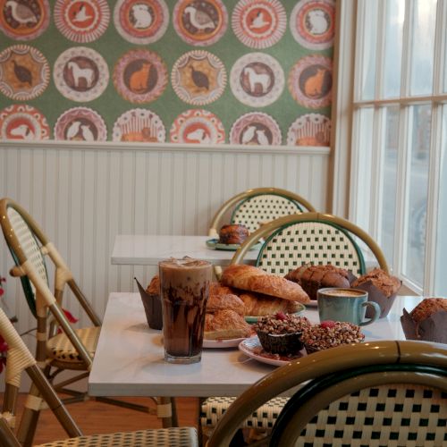 A cozy cafe scene with pastel wicker chairs, a table of pastries, coffee, and toast beside a window, in front of patterned wallpaper.