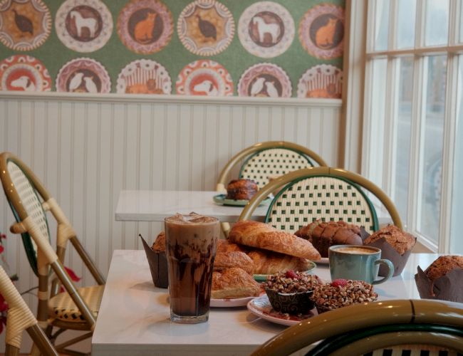 A cozy cafe scene with pastel wicker chairs, a table of pastries, coffee, and toast beside a window, in front of patterned wallpaper.