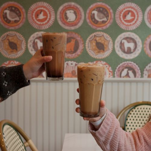Two people clink large iced coffees in a cozy caf&eacute;, raising toasting drinks in front of patterned retro wallpaper and wicker chairs.