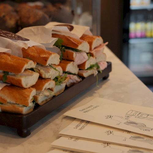 A tray of stacked club sandwiches with turkey, lettuce, and tomato on crusty bread, plus white napkins and takeaway orders on the counter.
