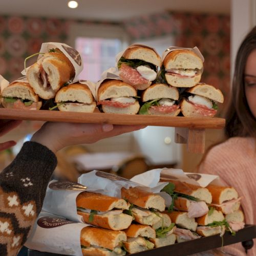 A person carries two trays of assorted mini sandwiches and wraps stacked high, ready for sharing.